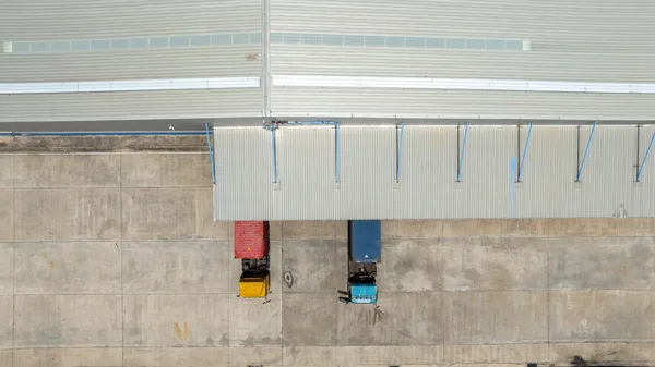 Vista aérea de camiones de carga, uno rojo y uno azul, posicionados debajo de un cobertizo junto a rampas hidráulicas.