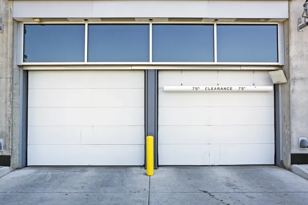 Dos puertas industriales de garaje blancas cerradas, con un poste amarillo en el medio y una señal de "7'0" CLEARANCE"