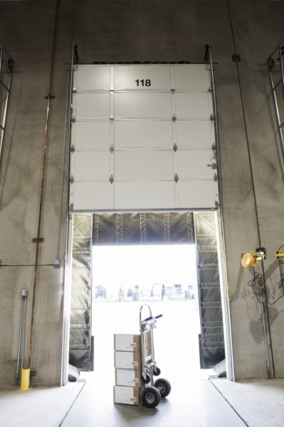 Puerta rápida blanca en muelle de carga con número 118 y carretilla con cajas frente a la salida abierta.