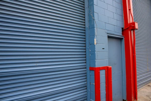 Una fachada de edificio de lamas metálicas azul grisáceo con detalles rojos, donde se distingue claramente una robusta cortina enrollable cerrada.